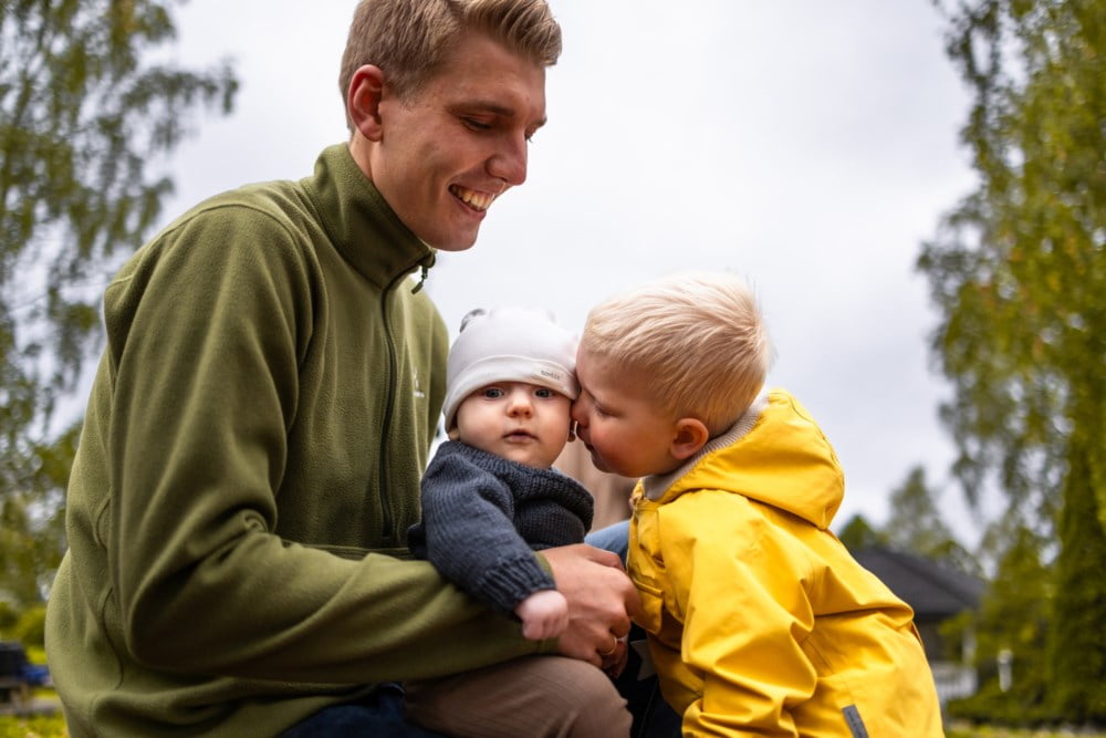 Far med veslebror på fanget sitt og storebror gir han ein klem