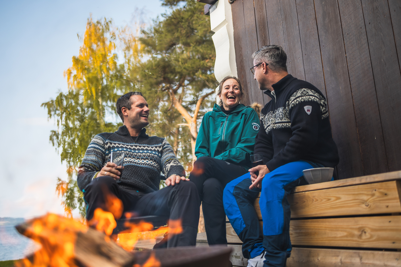 Tre menneske sit i hytteveggen framfor eit bål